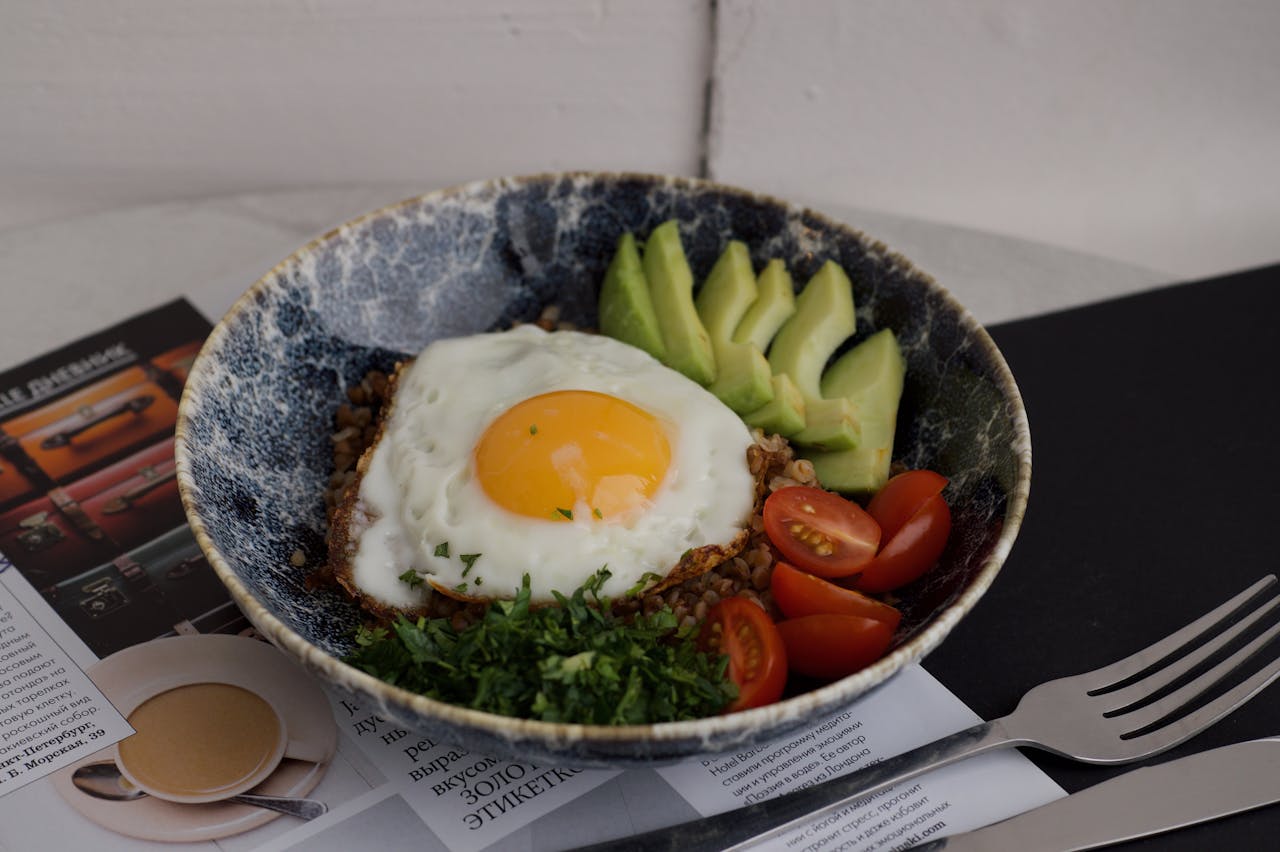 A nutritious breakfast bowl with a fried egg, avocado, tomatoes, and greens in a rustic setting.